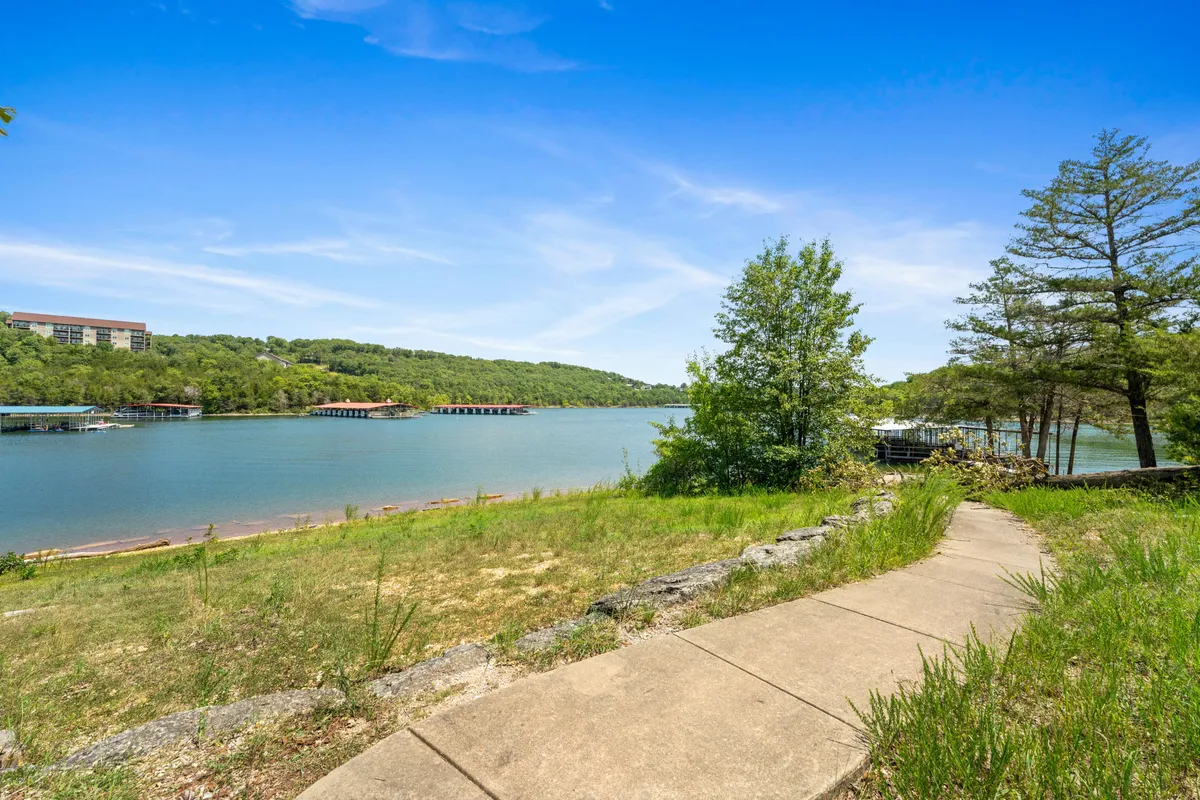 A serene waterfront view featuring a well-defined pathway winding along the lake, surrounded by lush greenery and distant hills under a tranquil sky. The area invites leisurely walks and peaceful moments by the water, offering ample space for outdoor exploration.