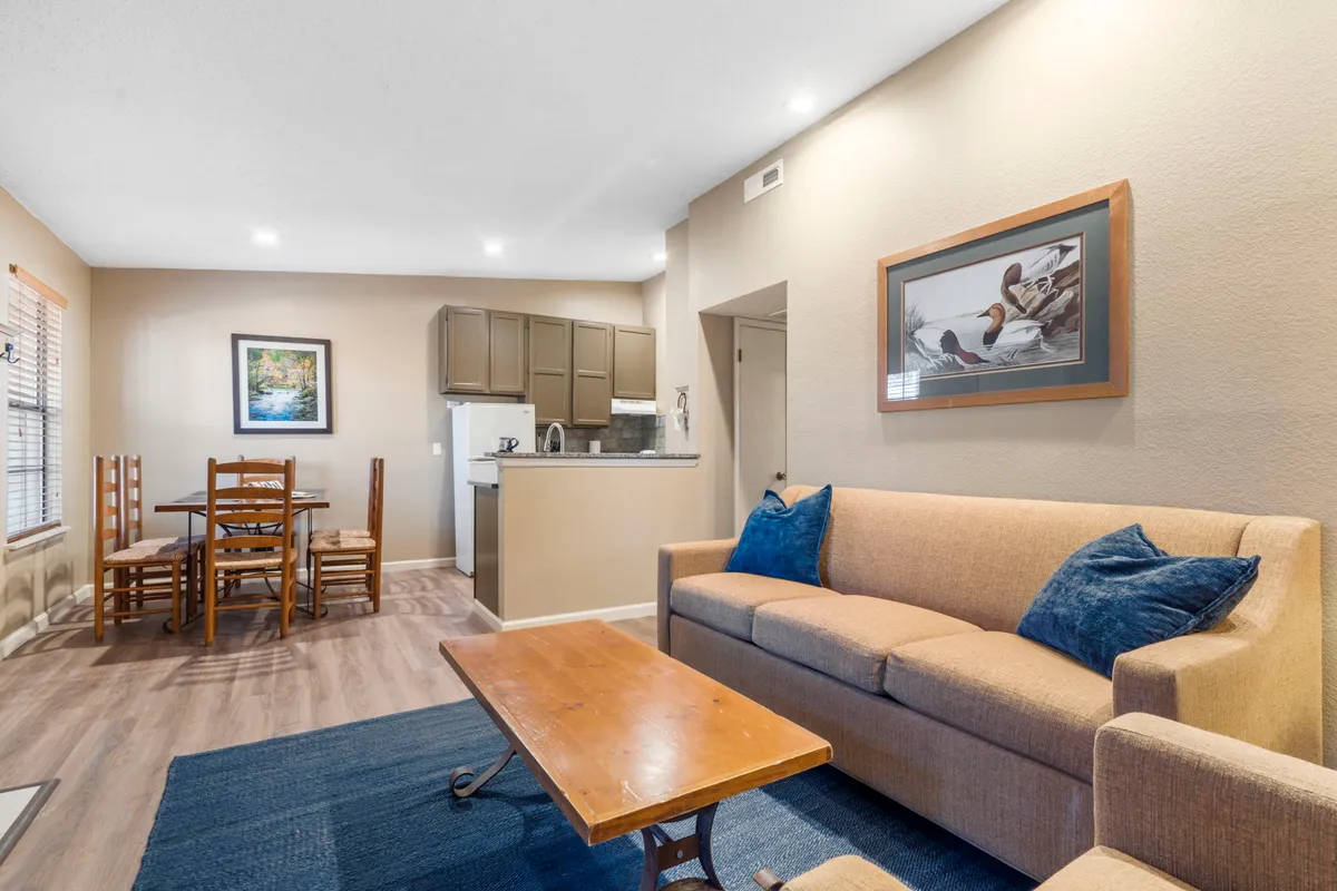 A bright interior scene with a comfortable sofa, a dining table ready for shared meals or games, and the kitchen area visible in the background. Artwork on the walls and tasteful décor create a warm, inviting setting ideal for socializing and relaxation.