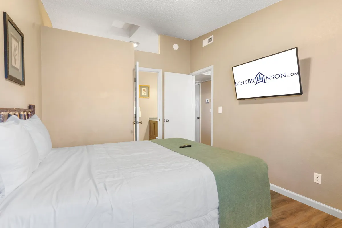 A comfortable bedroom with a plush queen bed dressed in soft linens, a wall-mounted TV, and doorways leading to an ensuite bath or other private spaces. The mood is tranquil and cozy, suited for relaxation after a busy day. Soft lighting and thoughtful décor contribute to a peaceful retreat.