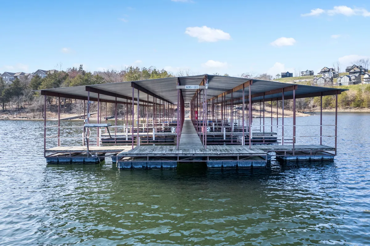 The focal point of this image is a dock extending into the lake, bordered by surrounding trees and houses on a gentle hill. The water’s surface is calm, perfect for fishing, launching boats, or simply relaxing by the lake. The setting is inviting and highlights the community's lakeside features.
