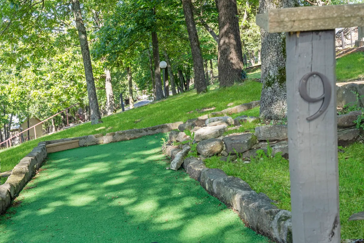 A vibrant mini-golf green nestled among mature trees, bordered with natural stone, and featuring a numbered post. The inviting setting includes space for groups to gather, play, and socialize, surrounded by lush greenery.