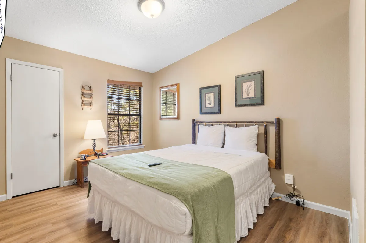 A serene bedroom scene featuring a queen bed, a bedside lamp, a window letting in gentle natural light, and subtle wall art. The space feels inviting, ideal for unwinding with a book or getting a restful night's sleep in quiet comfort.