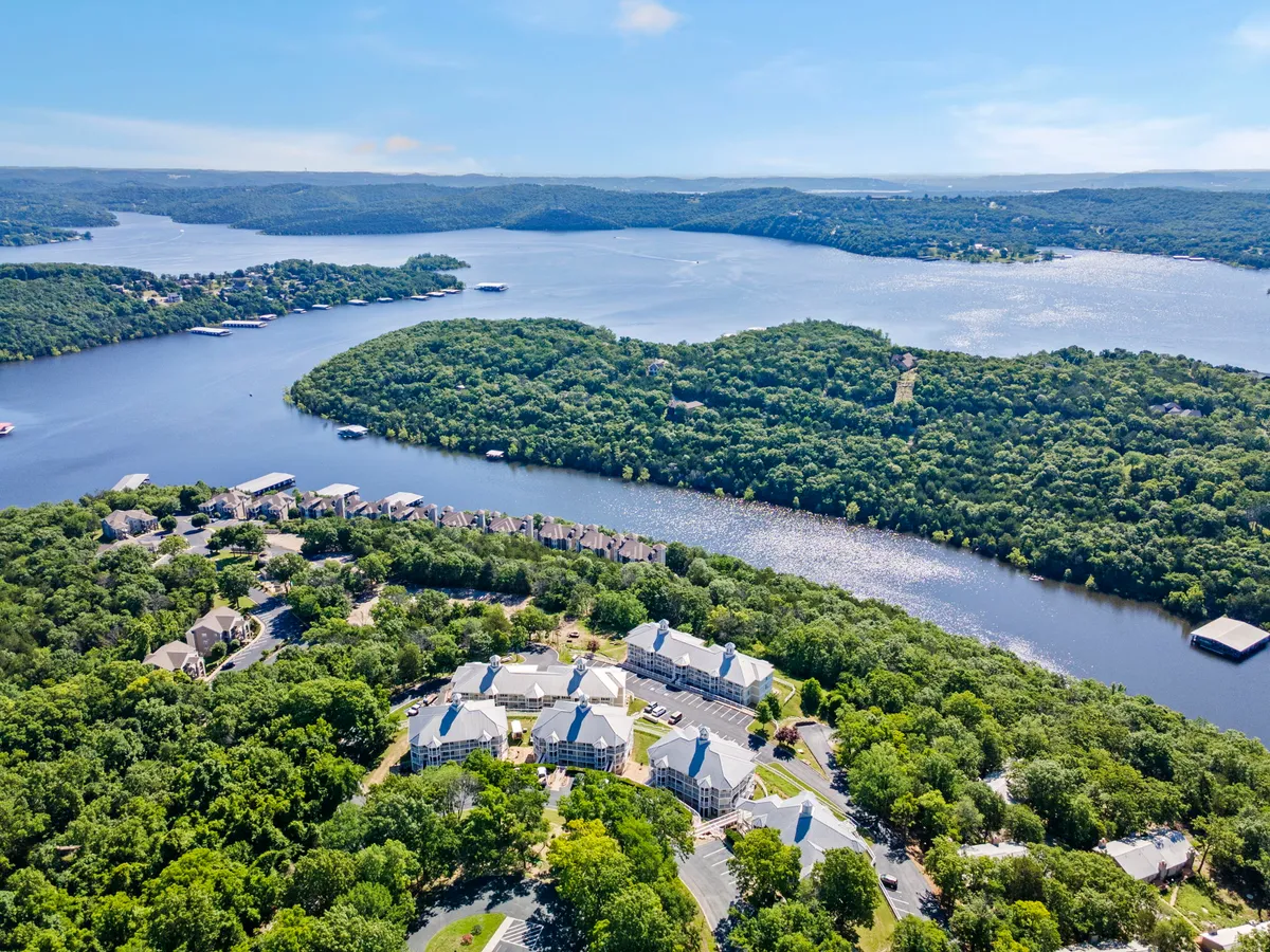 A wide lakeshore view, bordered by vibrant greenery and distant residential buildings tucked among the hills. The natural setting hints at walking trails and offers a sense of space ideal for both play and relaxation.